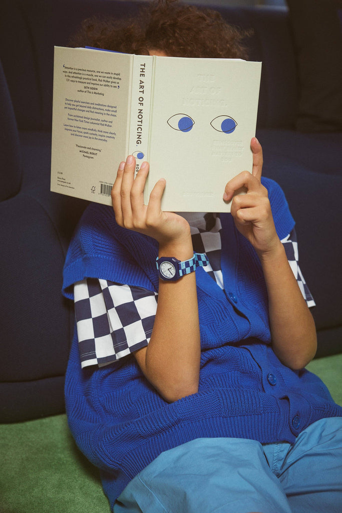 A child sits on a green carpet, wearing a blue vest and holding “The Art of Noticing,” while sporting the Mini Kyomo Watch - Blue Sunday - Blue Checkered on their wrist.