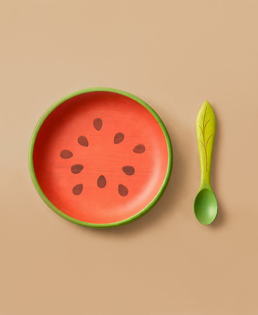 The Wally the Watermelon Plate & Spoon Set features a round, non-toxic plate painted like a watermelon slice and a green leaf-designed spoon—perfect stylish baby tableware on a tan background.