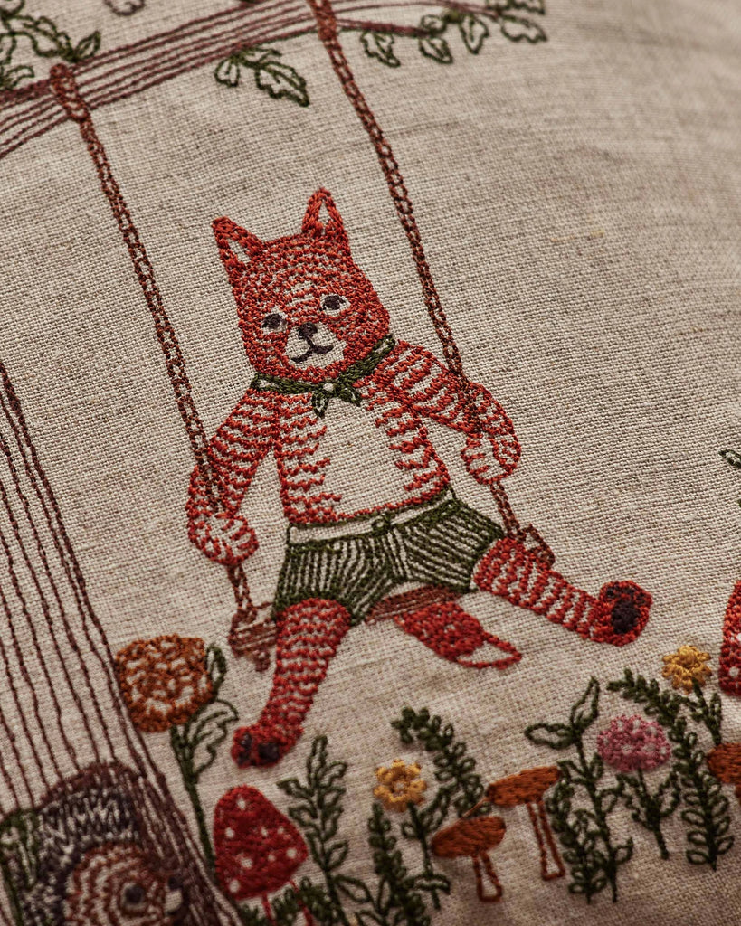 The Coral & Tusk Tree of Fun Pillow features an embroidered red-striped cat in green shorts on a swing, surrounded by flowers, mushrooms, and plants, bringing a playful touch to beige fabric.