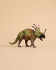 A small, green and brown Schleich Styracosaurus dinosaur toy with three horns and a frilled neck stands on a plain beige background.