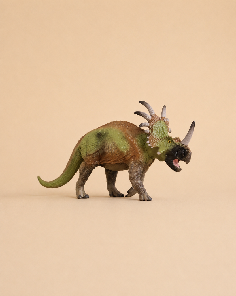 A small, green and brown Schleich Styracosaurus dinosaur toy with three horns and a frilled neck stands on a plain beige background.