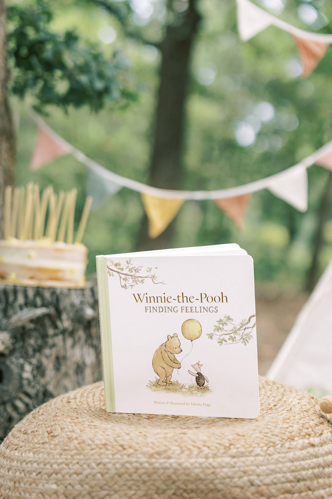 A “Winnie-the-Pooh: Finding Feelings” board book is displayed on a woven surface outside, with a blurred cake and pastel bunting visible in the background.