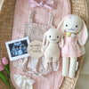 A baby announcement display featuring Cuddle + Kind Clover the Bunny (Blossom), a pink outfit, two stuffed bunnies, a wooden "My First Easter" sign, ultrasound photo, pink tulips, and white bloomers in a woven basket.