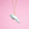 A gold chain necklace is shown against a pink background. The pendant is a small, handcrafted porcelain whale with a subtle glossy finish and a tiny black eye. The necklace appears to be suspended, creating an oceanic elegance for this whimsical White Beluga Necklace.