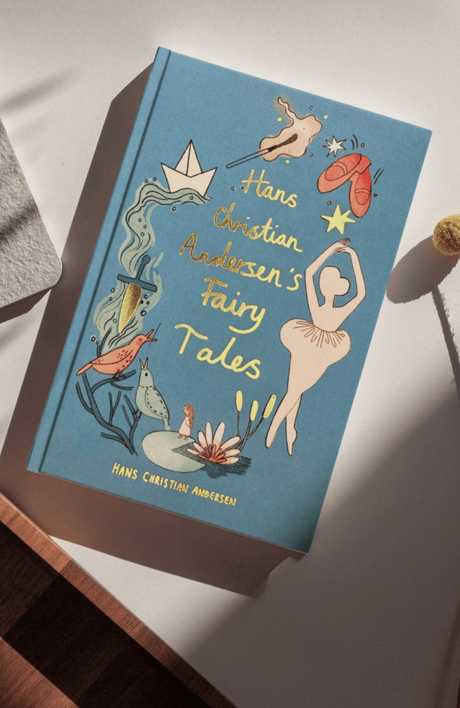 The "Hans Christian Andersen Fairy Tales | Collector's Edition," an illustrated blue book with whimsical cover art of a ballerina, paper boat, mermaid, birds, and stars, rests on a sunlit light surface.