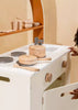 A child opens the door of a wooden play kitchen, discovering the Wooden Pots and Pans Playset (6 Pcs), a delightful kids cooking set featuring wooden pots, pans, and utensils arranged neatly on stove burners. The warm, neutral colors create a cozy and inviting play space.
