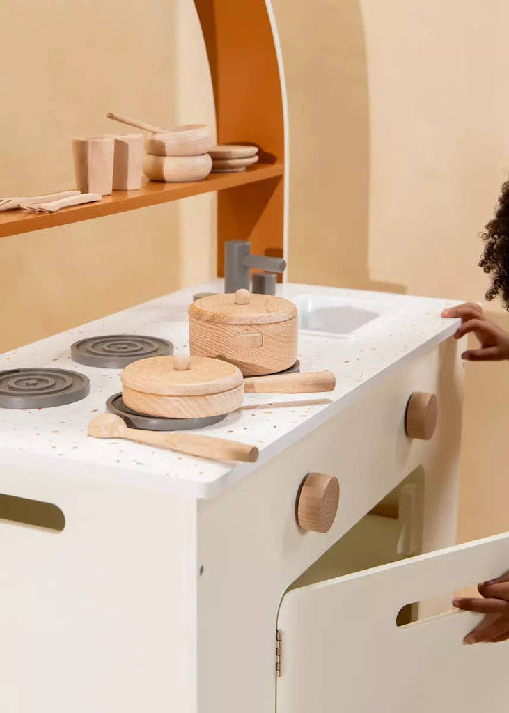 A child opens the door of a wooden play kitchen, discovering the Wooden Pots and Pans Playset (6 Pcs), a delightful kids cooking set featuring wooden pots, pans, and utensils arranged neatly on stove burners. The warm, neutral colors create a cozy and inviting play space.