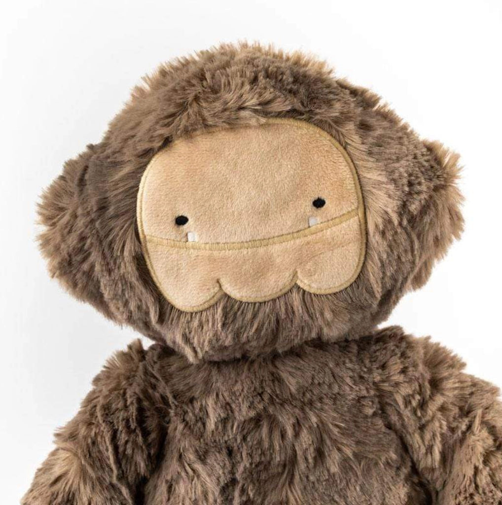 A close-up of a Slumberkins Bigfoot Kin plush toy with a beige face, small black eyes, and a smiling mouth line, filled with hypoallergenic fiberfill and covered in soft brown fur, set against