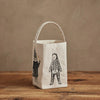 The Coral & Tusk Halloween Trick Or Treat Bag, a small cream bag with a handle and black line art of costumed critters, including a skeleton rabbit and a figure in a pointed hat, rests on a wooden surface against a beige wall. Ships in one week.