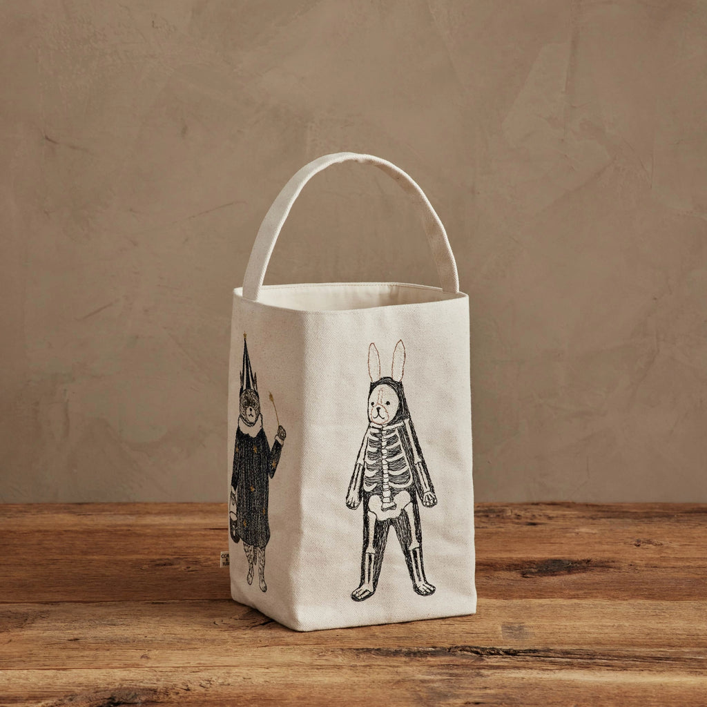 The Coral & Tusk Halloween Trick Or Treat Bag, a small cream bag with a handle and black line art of costumed critters, including a skeleton rabbit and a figure in a pointed hat, rests on a wooden surface against a beige wall. Ships in one week.