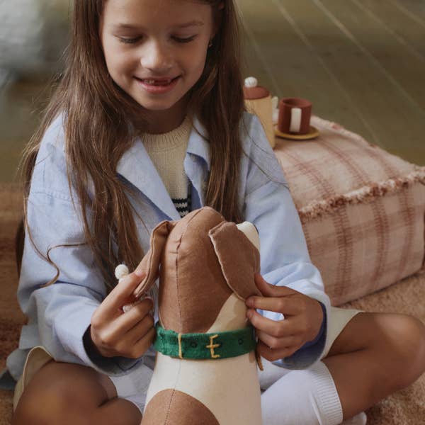 A young girl smiles while playing with the Dog Cushion - Cooper, a brown and white Beagle design with a green collar. She gently holds its ear, sitting on a patterned cushion with toy tea cups nearby—a charming handcrafted decor piece.