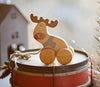 A Handmade Wooden Reindeer Pull Toy with wheels sits on a drum. The reindeer, made from sustainably sourced materials, has a red nose, a drawn-on scarf, and a cheerful expression. In the background, a small house-shaped ornament adds charm to the warmly lit scene.