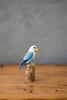 The Hand Carved Ornament - Lovebird - Light Blue - 7.5"H features a handcrafted blue and white lovebird with a yellow beak, perched on a short sustainable softwood stump, displayed on a wooden table with a plain gray background.