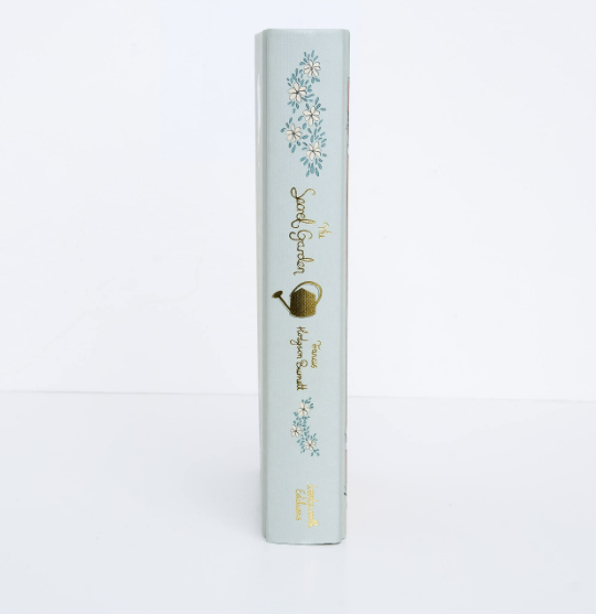 Spine of a light gray hardcover, "The Secret Garden," Collector's Edition by Burnett, adorned with decorative blue flowers and a gold watering can, reminiscent of Frances Hodgson Burnett's enchanting world.