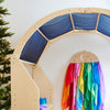 A wooden play structure with a gray fabric canopy invites imaginative play, while a rack of colorful Giant Playsilks, including Sarah's Silk Giant Midnight Playsilk, hangs nearby. A Christmas tree with lights is partially visible on the left.