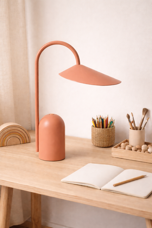 A minimalist wooden desk with the Ferm Living Arum Portable Lamp in Sienna, an open notebook and pencil, colored pencils in a basket, paintbrushes, wooden beads, and a rainbow-shaped decor piece.