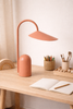 A minimalist wooden desk with the Ferm Living Arum Portable Lamp in Sienna, an open notebook and pencil, colored pencils in a basket, paintbrushes, wooden beads, and a rainbow-shaped decor piece.