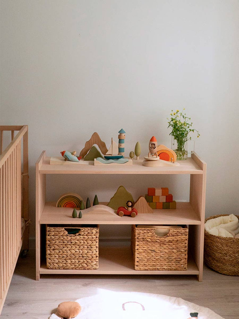 The Sabo Concept First Journey sits on a wooden shelf with a vase of flowers and two woven baskets in a softly lit, minimalist children’s room with a crib and basket nearby.