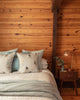 A cozy bedroom features a wood-paneled wall, a bed with the Coral & Tusk Downhill Skiers Pillow and patterned pillows, plus a textured blanket. A wooden nightstand holds a glass lamp, small plant, and a book.