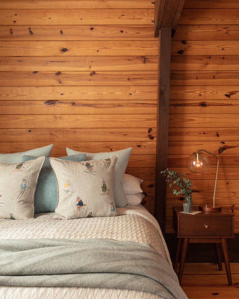 A cozy bedroom features a wood-paneled wall, a bed with the Coral & Tusk Downhill Skiers Pillow and patterned pillows, plus a textured blanket. A wooden nightstand holds a glass lamp, small plant, and a book.