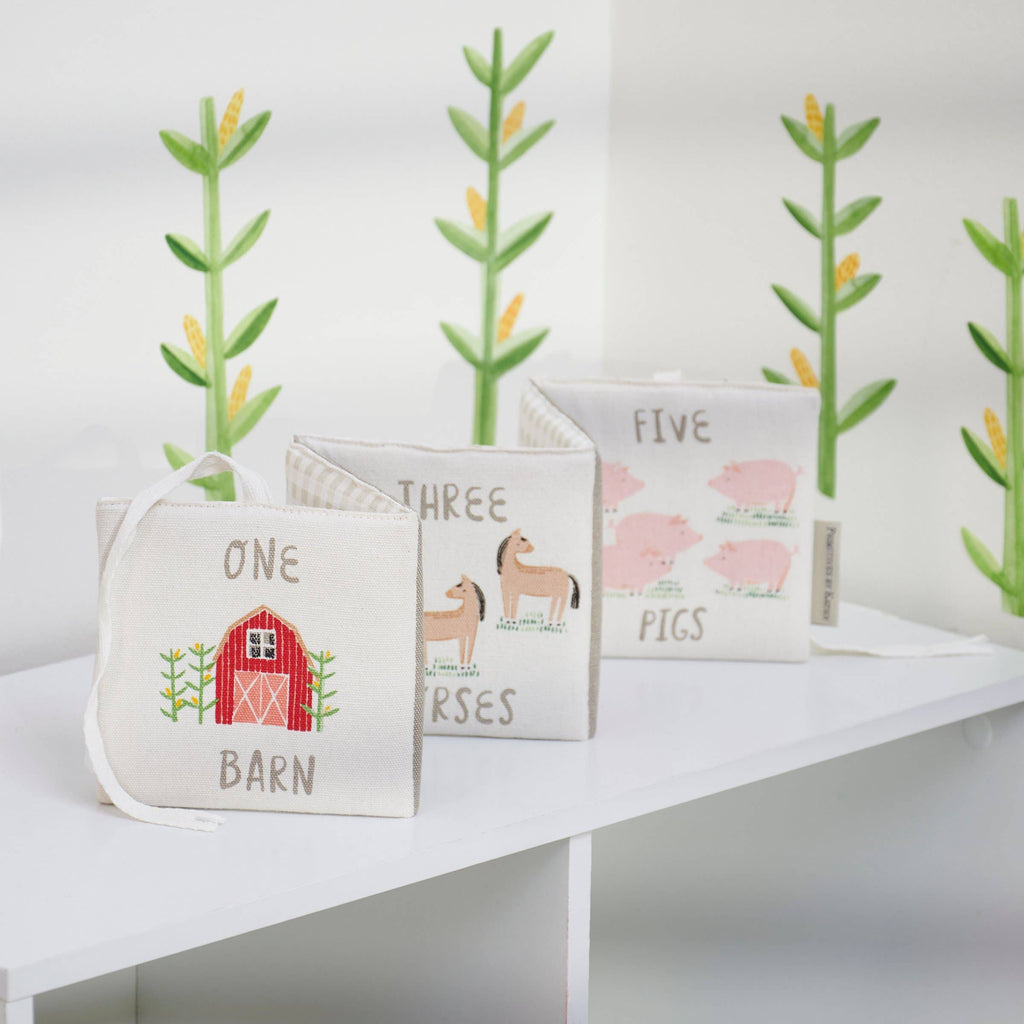 The Little Farm Soft Book set includes three embroidered books—“One Barn,” “Three Horses,” and “Five Pigs”—making a perfect farm-themed gift for infants. Displayed on a white shelf with plant decals in the background.
