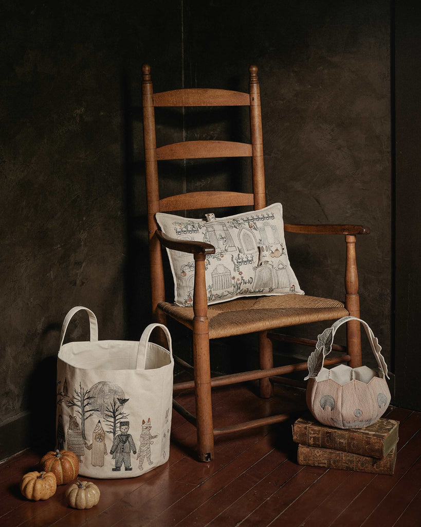 A wooden rocking chair with a Coral & Tusk Graveyard Pocket Pillow, a fabric basket with spooky designs, stacked books, small pumpkins, and a unique basket on a dark wooden floor against a dark wall.