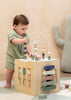 A toddler in a green outfit plays with the Wooden Activity Cube, featuring colorful beads, spinning blocks, and xylophone bars, in a cozy room with neutral decor. This classic toy supports fine motor skill development.