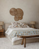 A cozy bedroom with a neutral palette features a bed with nautical accent pillows, including the Coral & Tusk Sailing Foxes Pillow, rustic wooden side tables and bench, woven lamps, and wicker wall art above the headboard.