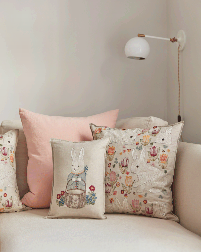 A beige sofa is decorated with four pillows, including the Coral & Tusk Easter Bunny Pocket Pillow featuring a bunny in a blue jacket, plus floral designs, creating a charming Easter look beneath a modern wall-mounted lamp.