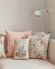 A beige sofa is decorated with four pillows, including the Coral & Tusk Easter Bunny Pocket Pillow featuring a bunny in a blue jacket, plus floral designs, creating a charming Easter look beneath a modern wall-mounted lamp.