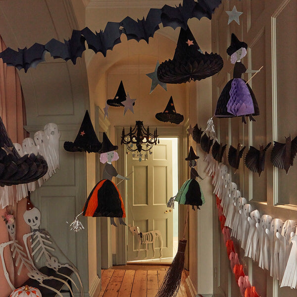 A Halloween party decorates the hallway with hanging witch hats, flying bats, and ghost garlands. Skeleton figures sit on a bench to the left, while Meri Meri Flying Honeycomb Witch Decorations float eerily above. A small skeleton dog stands in a lit room through an open door at the end of the hallway.