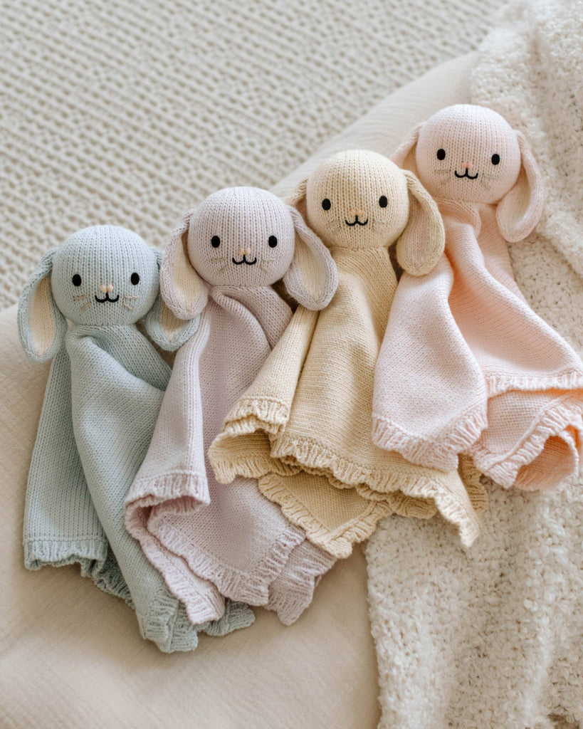 The Cuddle + Kind Organic Cotton Baby Bunny Lovey in Baby Blue, with floppy ears and a smiling face, is hand-knit and soft—perfect for cuddling on a cream-colored textured blanket.