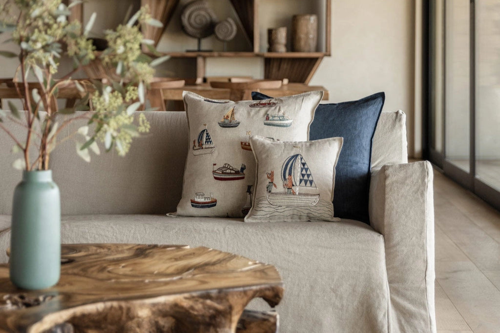A beige sofa with a Coral & Tusk Sailing Foxes Pillow featuring boat and sailor motifs brings a nautical touch beside a wooden coffee table. In the foreground, a green vase with foliage stands out against shelves and decor in the background.