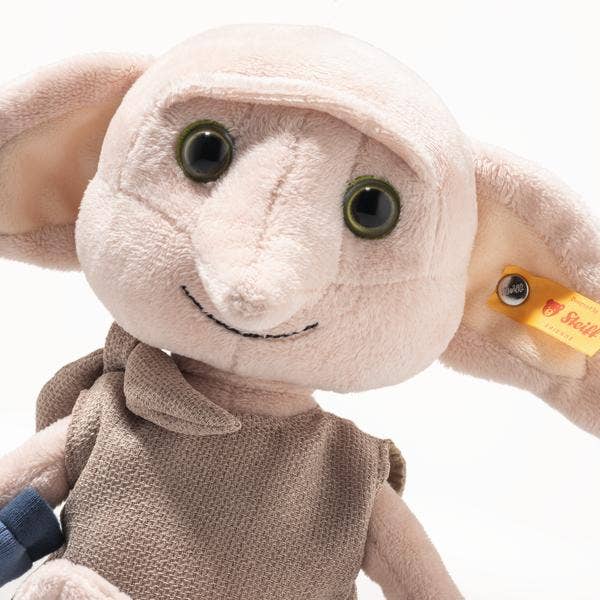 The Steiff Harry Potter “Dobby” with Sock 10" plush toy features large floppy ears, green eyes, a beige tunic, a yellow Steiff tag on one ear, and is displayed against a white background.
