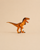 A small, orange Schleich Velociraptor dinosaur toy with black stripes stands on a beige background, mouth open and arms raised, featuring a movable jaw for added playability.