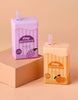 The Tiny Tummies Set - Juice Box Duo includes two interactive toy juice boxes—one grape and one orange—each with a straw. Featuring pastel colors, these doll accessories are displayed on a tan background atop a raised block.