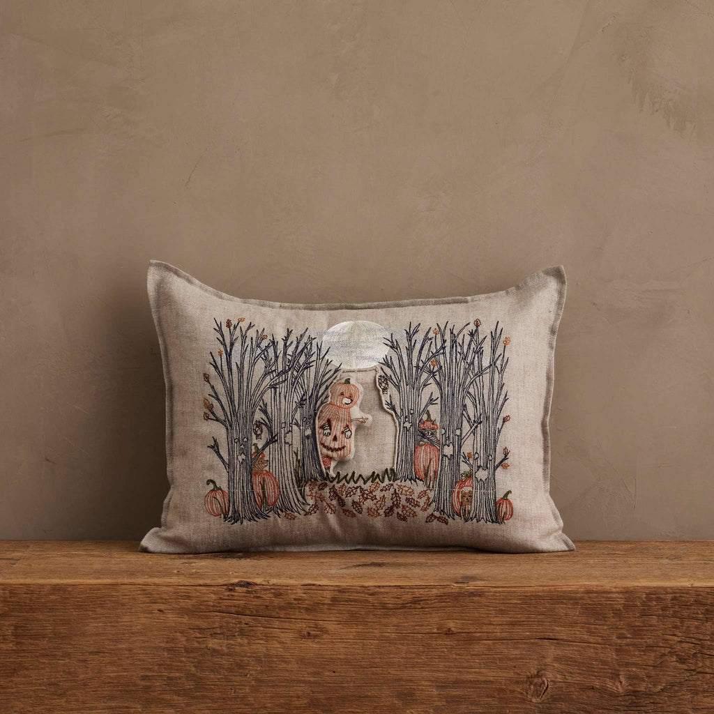 A Coral & Tusk Spooky Forest Pocket Pillow featuring embroidered bare trees, pumpkins, and a central ghostly skull face sits against a plain wall on a wooden surface.