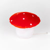 The Red Mushroom Lamp features a bright red and white spotted cap with a round white base and plugs in with a white cord—ideal as a whimsical night light for nurseries or children’s rooms.