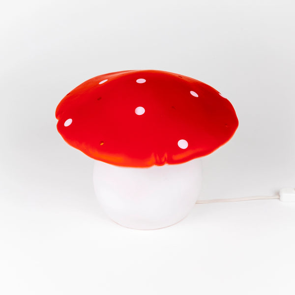 The Red Mushroom Lamp features a bright red and white spotted cap with a round white base and plugs in with a white cord—ideal as a whimsical night light for nurseries or children’s rooms.
