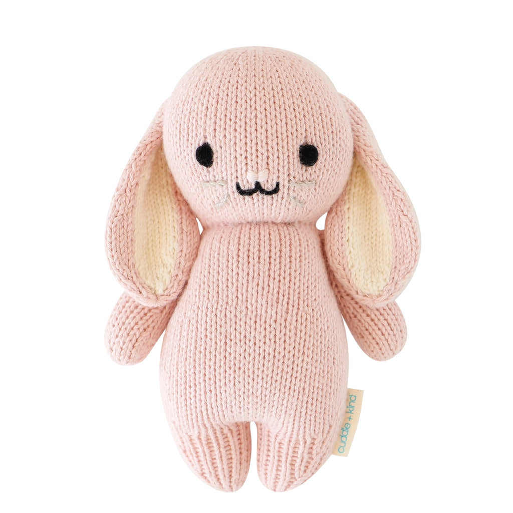 The Cuddle + Kind Baby Bunny (Rose) is a soft, heirloom-quality plush toy, hand-knit from Peruvian cotton yarn with long floppy ears, black stitched eyes, a sweet smile, and a side tag.