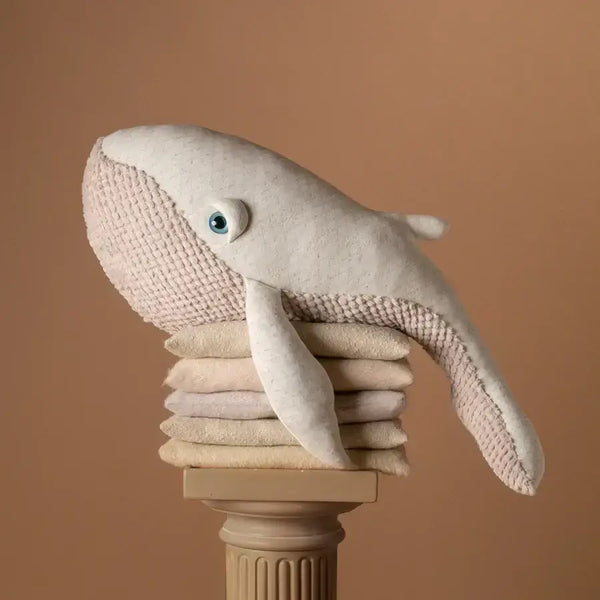 A BigStuffed The Big Whale Albino stuffed animal with a textured surface sits atop folded towels on a decorative column, ready for ocean adventures against a neutral beige background.