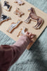 A child’s hand places a piece from the Farm Animals Wooden Puzzle: A Tabitha Paige Puzzle onto its board, featuring beautifully painted farm animals and resting on a textured fabric surface—an engaging wooden peg puzzle and the perfect educational toy for toddlers.