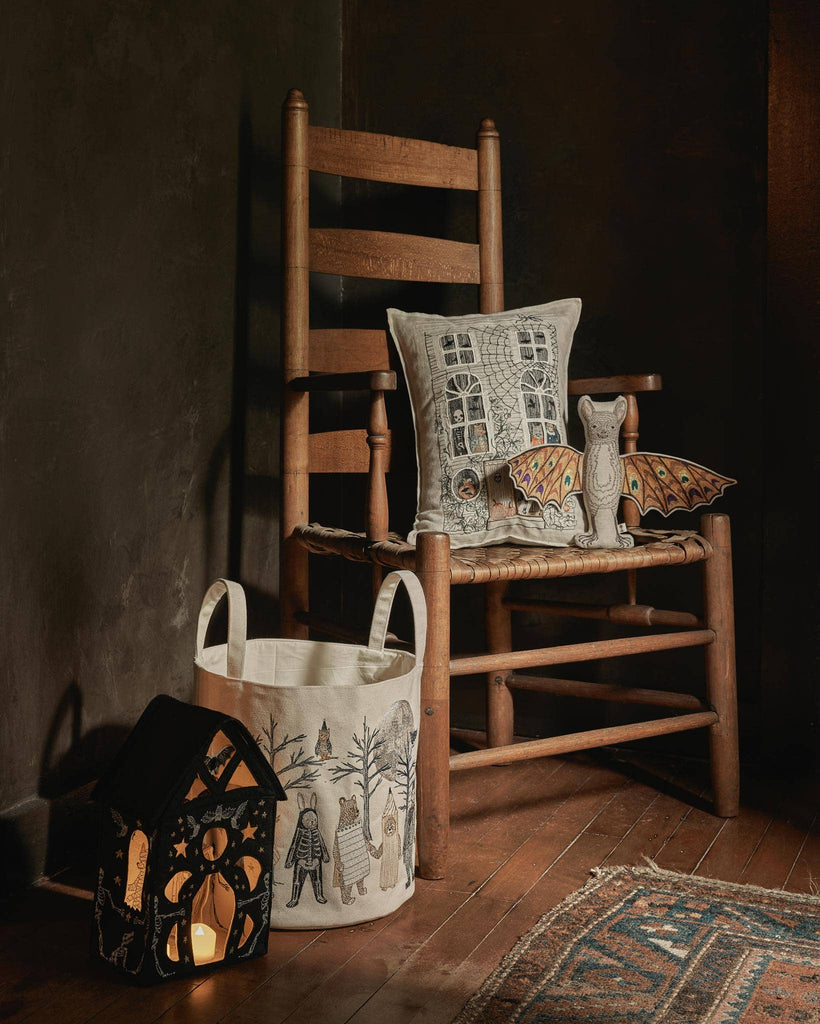 A Coral & Tusk Bat Surprise Doll with fabric bat wings rests on a wooden rocking chair with a decorative pillow, beside a winter-themed storage bin and a glowing black house-shaped lantern. Ships in one week.