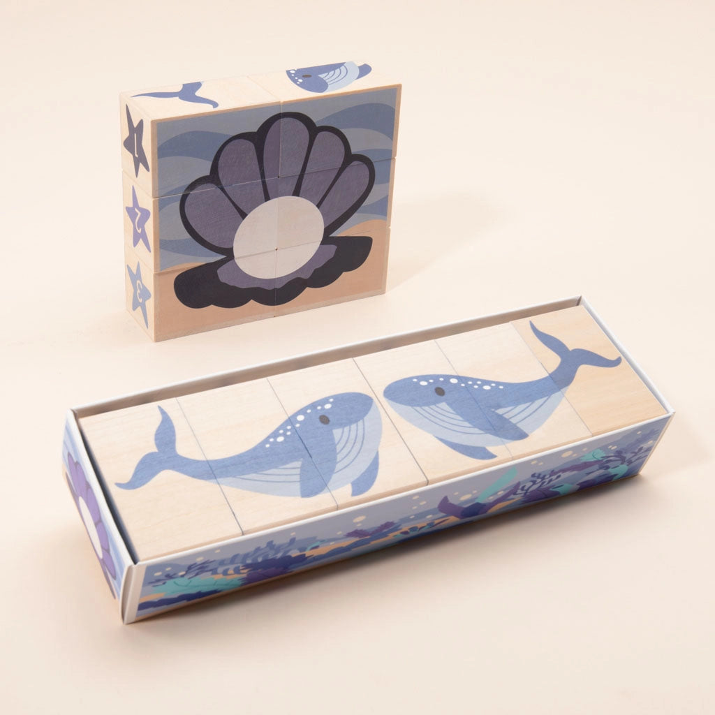 A set of Ocean Blocks is shown, arranged in two separate configurations. The rectangular arrangement in the foreground depicts blue whales, while the background displays a shell with a pearl. Both sets, made from sustainable basswood, are illustrated in shades of blue.