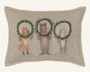 The Christmas Wreath Trio Pocket Pillow features a fox, bear, and rabbit standing upright on a beige background, each holding a holiday wreath decor in front of their faces.