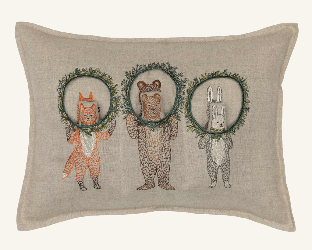 The Christmas Wreath Trio Pocket Pillow features a fox, bear, and rabbit standing upright on a beige background, each holding a holiday wreath decor in front of their faces.