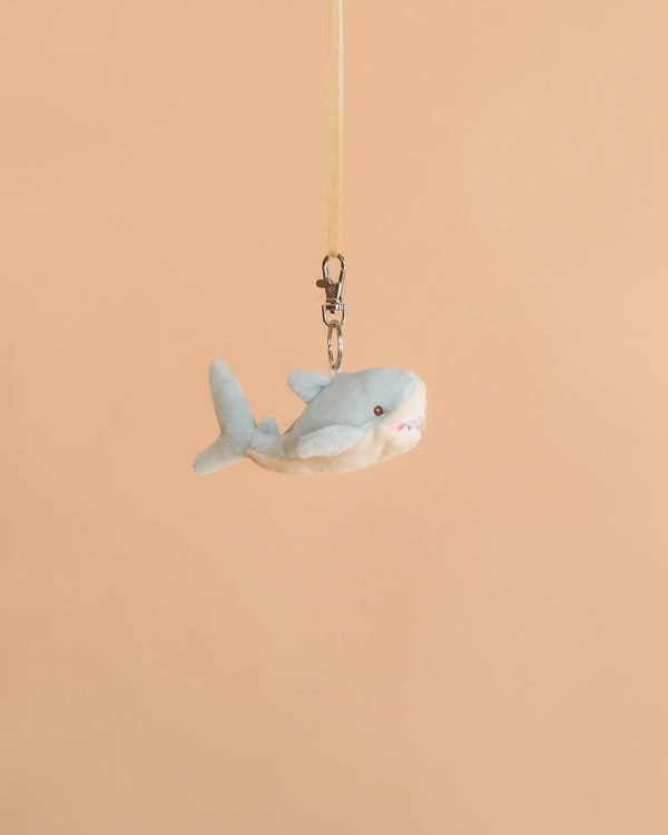 The Bukowski - Splash Shark Bag Charm, a small plush shark keychain perfect for cuddling, hangs from a beige ribbon against a plain peach background.