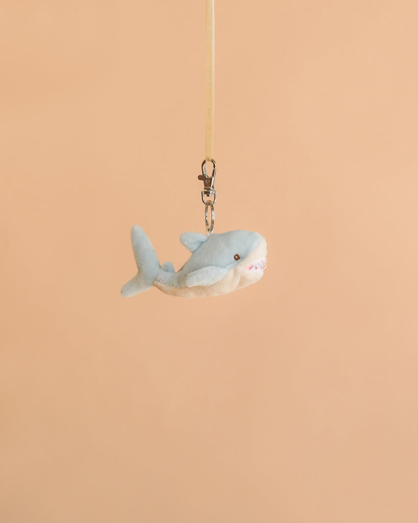 The Bukowski - Splash Shark Bag Charm, a small plush shark keychain perfect for cuddling, hangs from a beige ribbon against a plain peach background.
