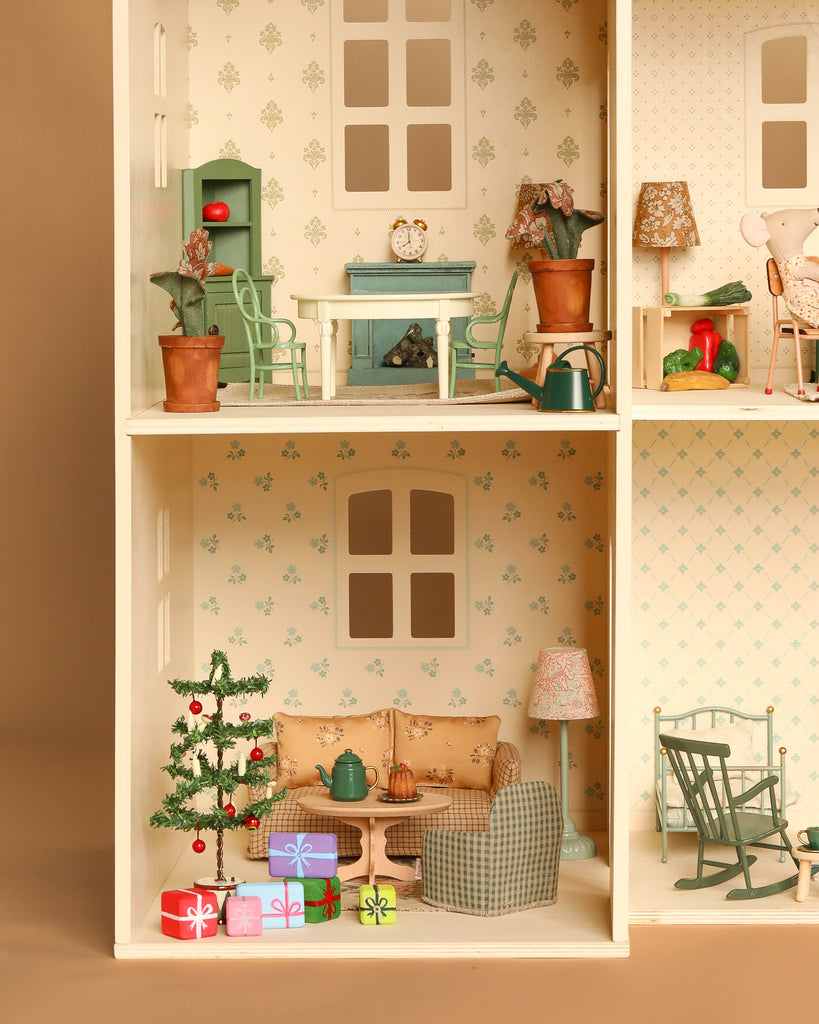 The Maileg Fully Furnished Holiday Dollhouse Set includes floral wallpaper, dollhouse furniture, a mini Christmas tree with ornaments, wrapped gifts, sofa, armchair, rocking chair, lamp, and coffee table with cups and pastries.