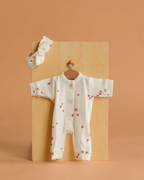 The Minikane 11" Doll Clothing set, featuring the Lili Jumpsuit & Headband in Ribbed Knit - Pink Hearts, is displayed on a wooden board against a light brown background, perfect as stylish doll clothes.